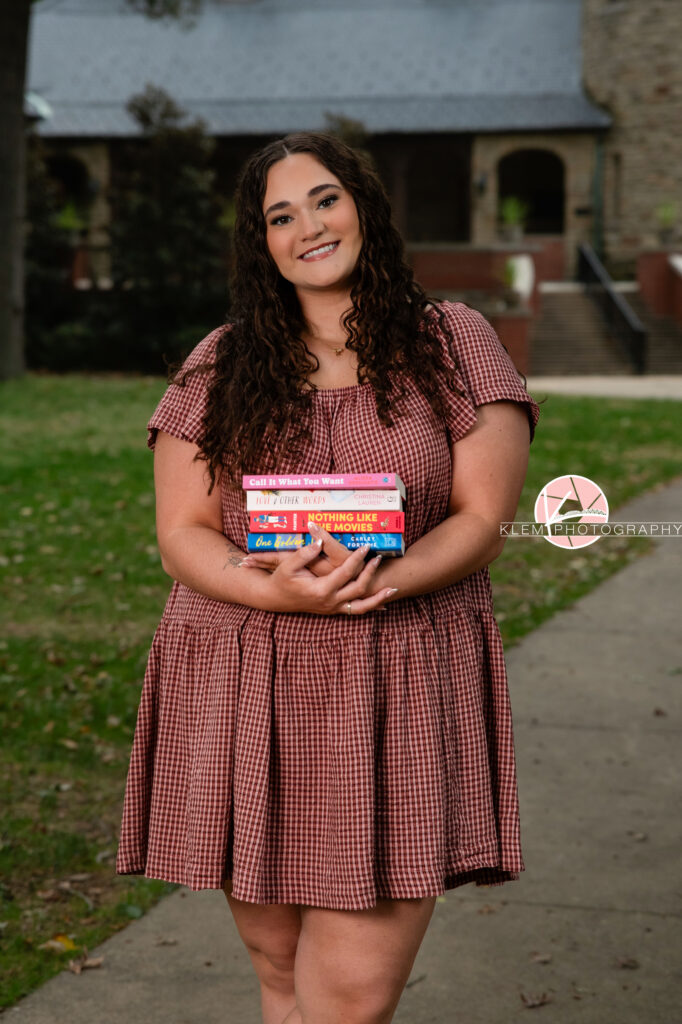 Henderson KY Senior Picture Ideas, Klem Photography, Mackenzie, Henderson County High School. Half body shot of senior girl with long brown curly hair wearing a red gingham dress smiles at the camera. She stands holding her books in front of her with a castle in the background.