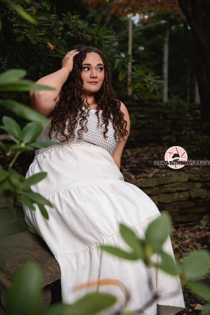 Henderson KY Senior Picture Ideas, Klem Photography, Mackenzie, Henderson County High School. Full body shot of senior girl with long brown curly hair wearing a white dress looks off camera. She sits on a stone bench with one hand in her hair with stone bricks and greenery in the background