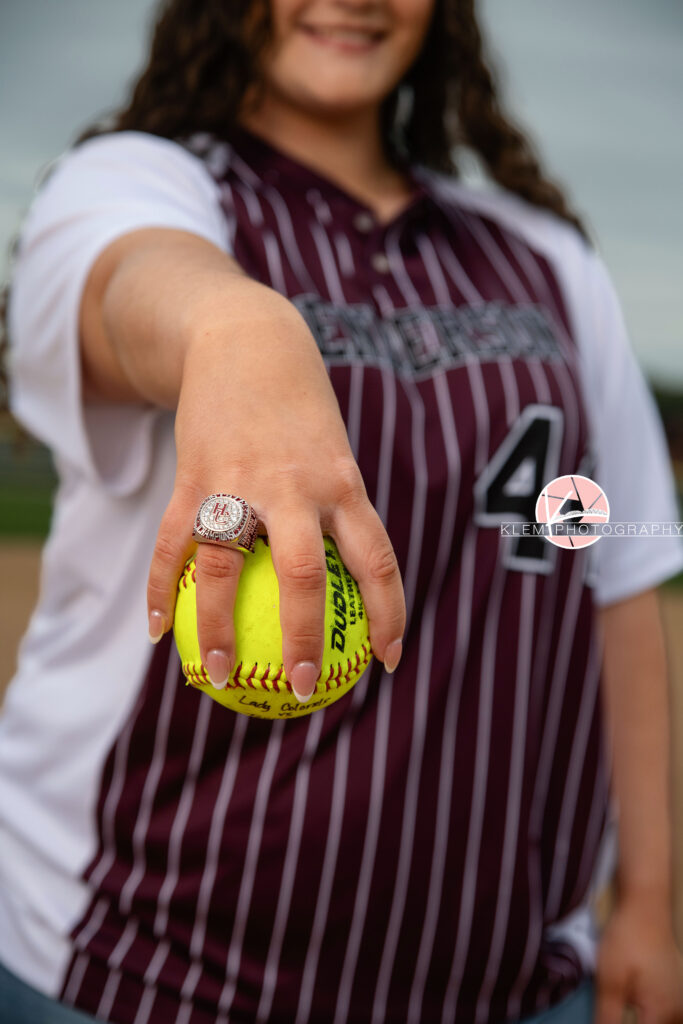 Henderson KY Senior Picture Ideas, Klem Photography, Mackenzie, Henderson County High School. Mid body shot of senior girl with long brown curly hair wearing a softball jersey smiles at the camera. She stands holding a softball in focus of the camera with her class ring on display with her other hand at her side.