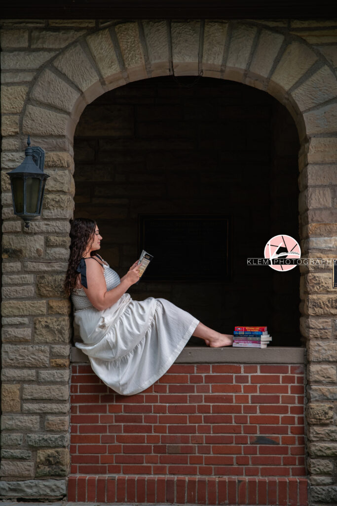 Henderson KY Senior Picture Ideas, Klem Photography, Mackenzie, Henderson County High School. Full body shot of senior girl with long brown curly hair wearing a white dress reading a book. She sits on a red brick wall under a stone brick arch holding a book with more books stacked by her feet.