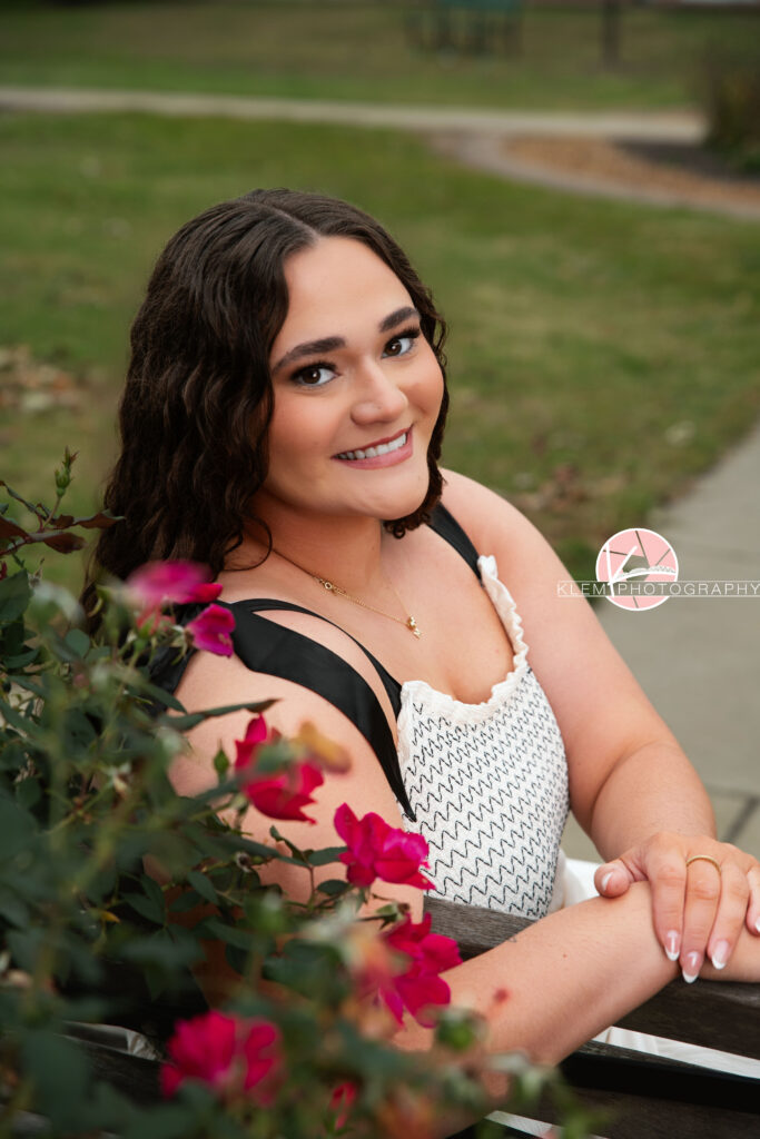 Henderson KY Senior Picture Ideas, Klem Photography, Mackenzie, Henderson County High School. Half body shot of senior girl with long brown curly hair wearing a white dress smiles at the camera. She sits with her hands on top of one another with a rose bush in the foreground and grass in the background.