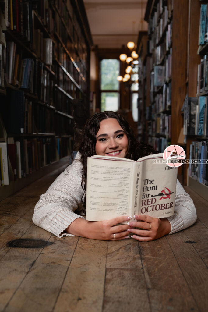 Henderson KY Senior Picture Ideas, Klem Photography, Mackenzie, Henderson County High School. Front shoulder shot of senior girl with long brown curly hair wearing a white sweater smiles at the camera. Sher face is visible over a book as she lays on a wooden floor and holds a book in front of her. There are bookcases around her.