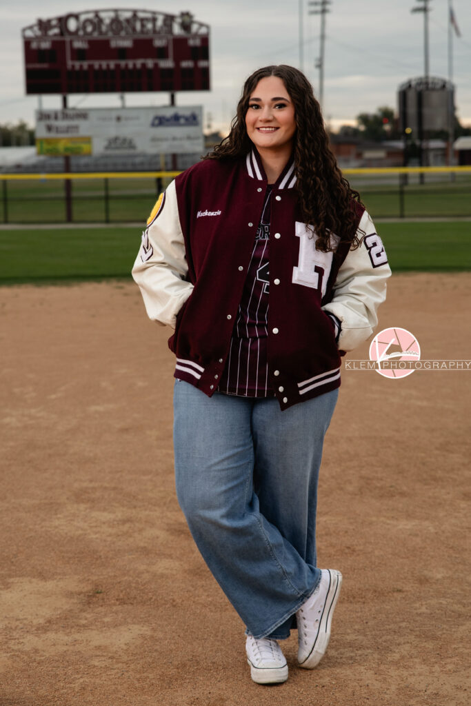 Henderson KY Senior Picture Ideas, Klem Photography, Mackenzie, Henderson County High School. Full body shot of senior girl with long brown curly hair wearing a softball jersey, letterman jacket, jeans, and white converse smiles at the camera. She stands with her hands in her pockets and one leg crossed in front of the other with a softball field in the background.