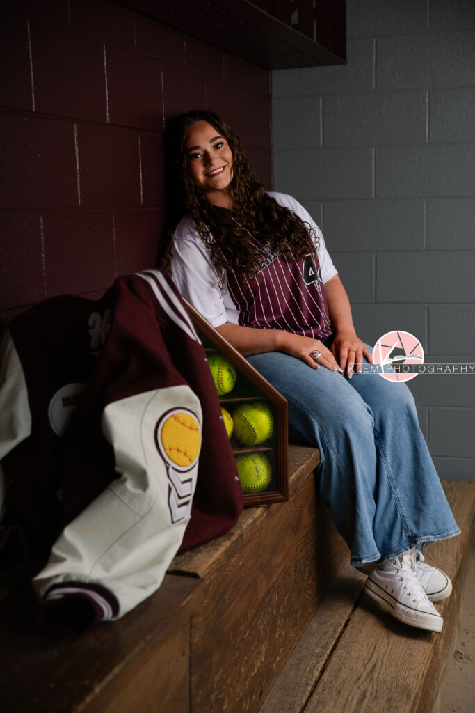 Henderson KY Senior Picture Ideas, Klem Photography, Mackenzie, Henderson County High School. Full body shot of senior girl with long brown curly hair wearing a softball jersey, jeans, and white converse smiles at the camera. She sits on a wooden bench with her legs crossed and her arms resting on her legs. Next to her is a container of softballs and a letterman jacket. There is a maroon and grey painted concrete brick wall in the background.
