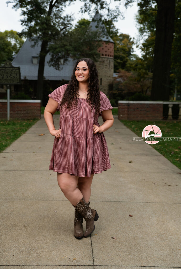 Henderson KY Senior Picture Ideas, Klem Photography, Mackenzie, Henderson County High School. Full body shot of senior girl with long brown curly hair wearing a red gingham dress and brown cowboy boots smiles at the camera. She stands with her hands on her hips and one leg crossed in front of the other. There is a castle and greenery in the background.